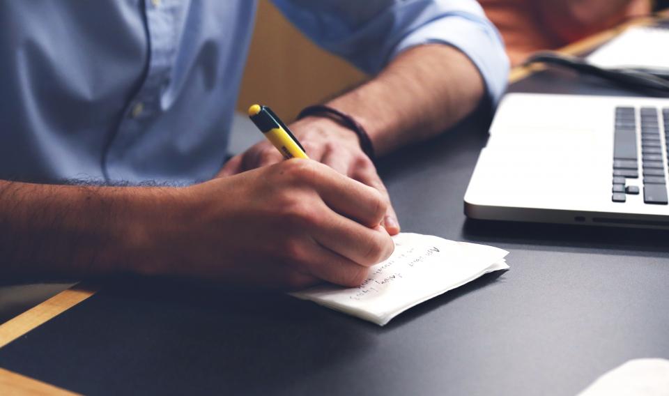 Person writing notes beside a laptop